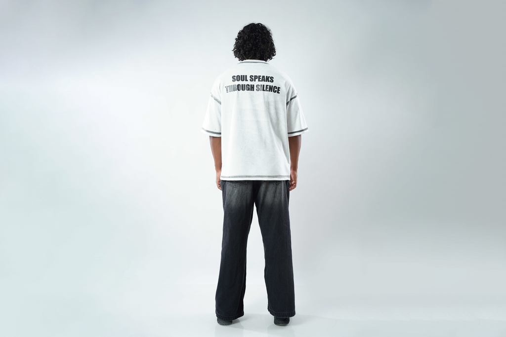 A person viewed from behind wearing a white short-sleeve T-shirt with black text reading 'SOUL SPEAKS THROUGHOUT SILENCE' on the back, paired with dark wide-leg pants, against a light gray studio background.