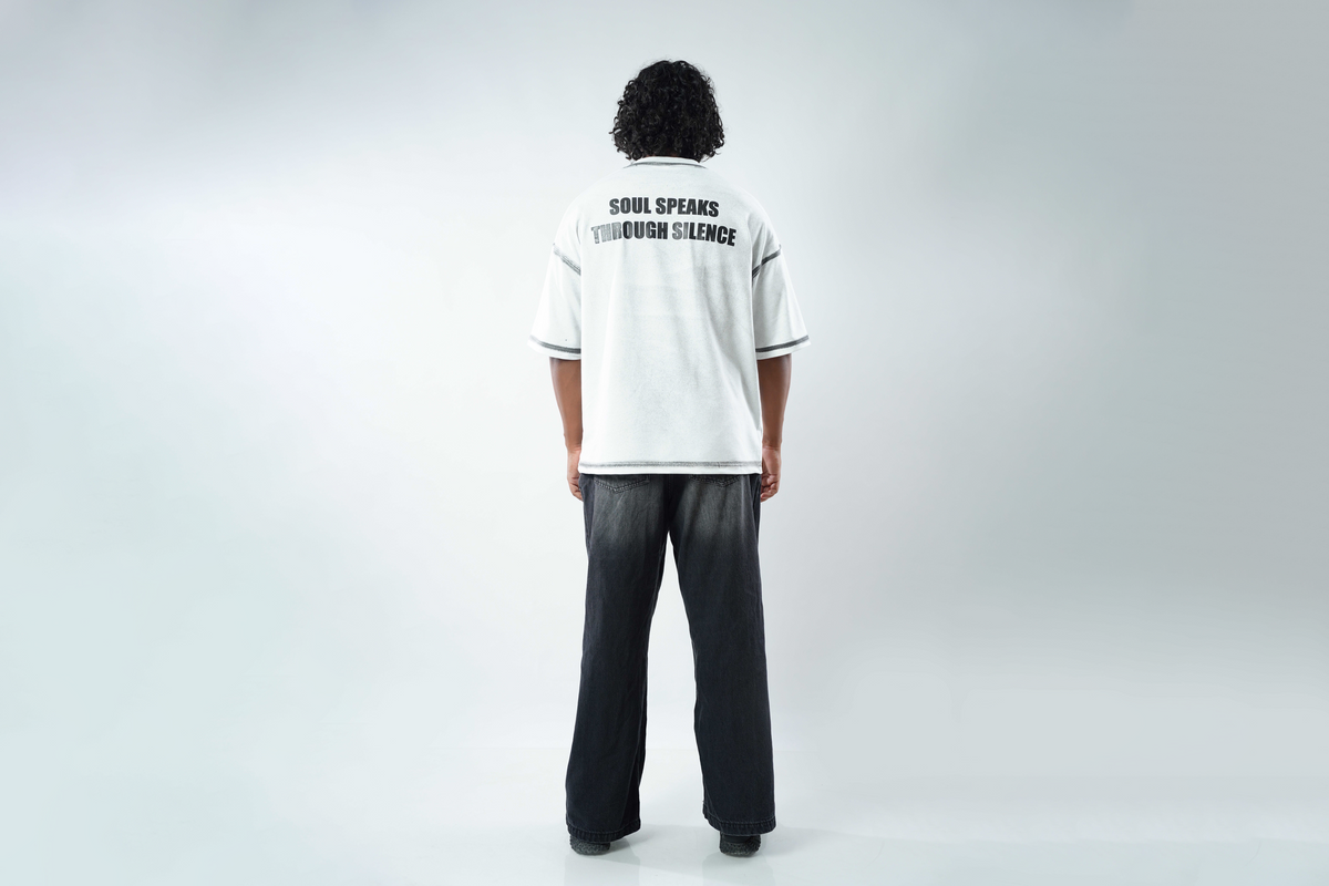 A person viewed from behind wearing a white short-sleeve T-shirt with black text reading 'SOUL SPEAKS THROUGHOUT SILENCE' on the back, paired with dark wide-leg pants, against a light gray studio background.