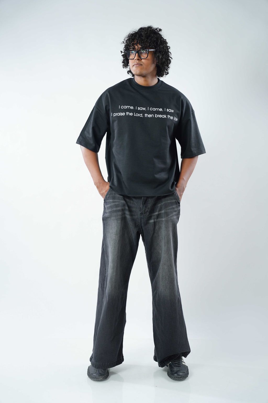 A man wearing a black graphic t-shirt with white text, dark gray wide-leg jeans, and black shoes, standing against a light gray studio background.