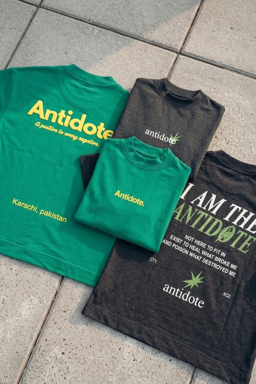 Four graphic t-shirts in green and dark gray are arranged in a fan shape on a gray tiled floor. The shirts feature the word 'Antidote' in yellow and white, with various text slogans and a small green leaf logo.